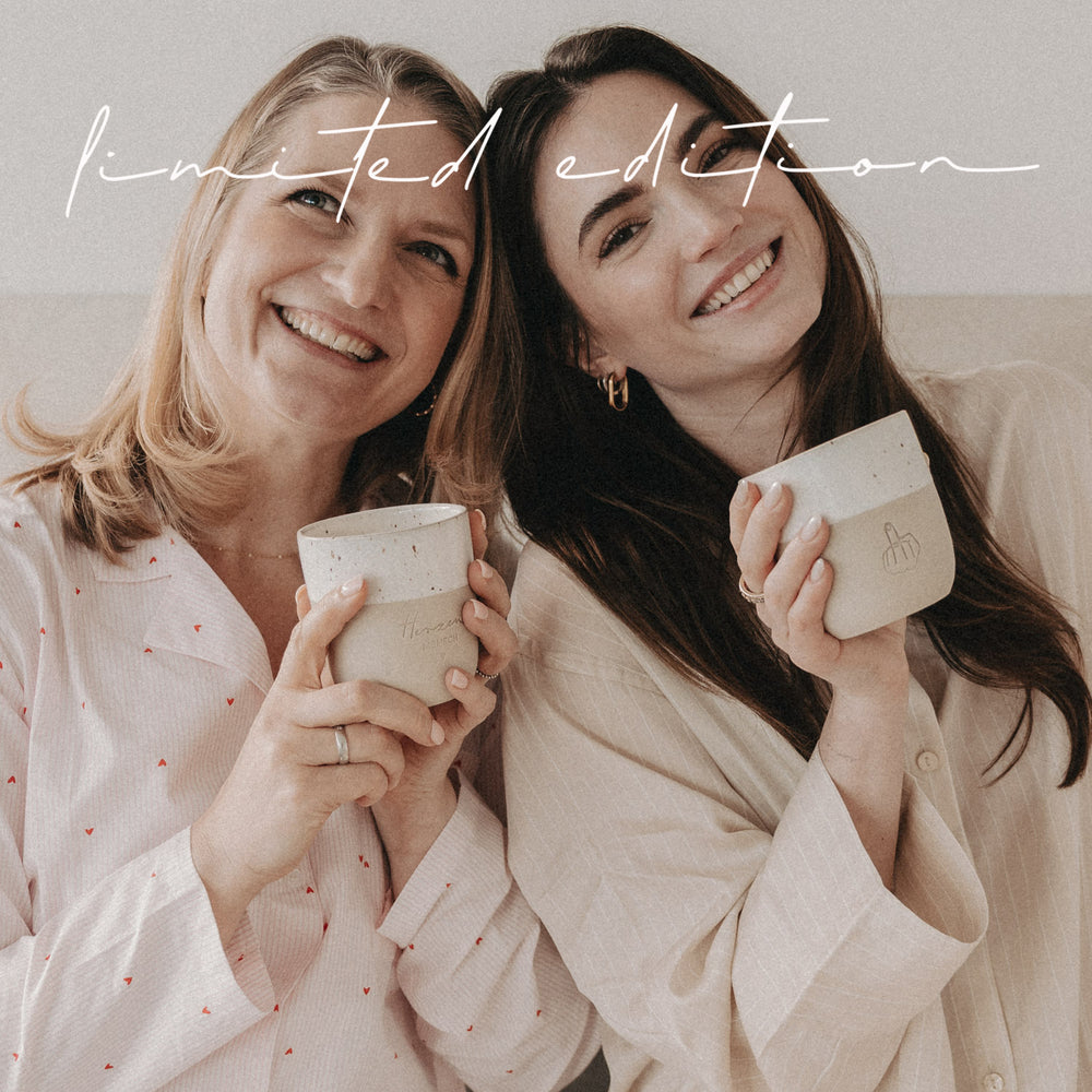 Frau hält beige-weißen Limited-Edition-Becher mit Mittelfinger-Motiv seitlich, lächelnd und in gemütlicher Umgebung.