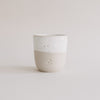 Keramikbecher in Beige und Weiß mit drei Herzprägungen, minimalistisch fotografiert vor hellem Hintergrund.