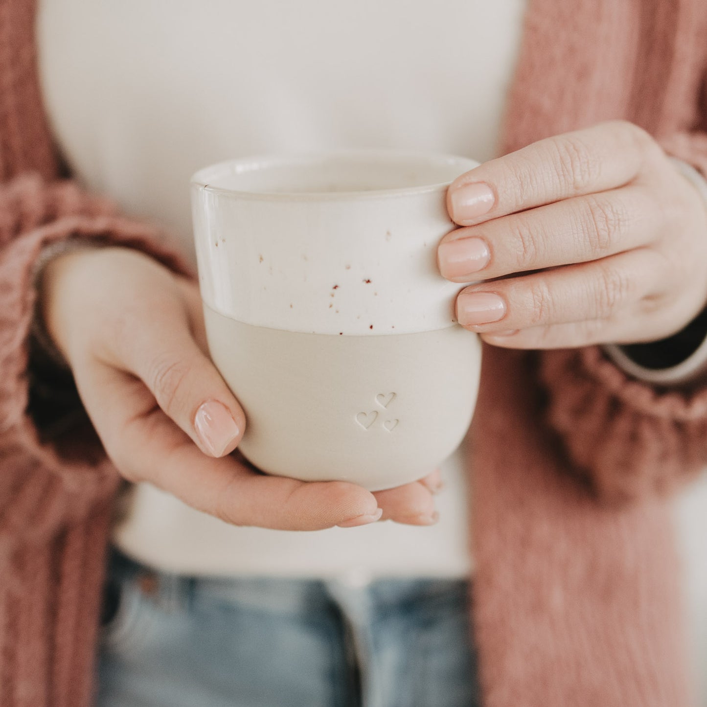 Hände halten den beige-weißen Keramikbecher mit drei Herzen; warme, gemütliche Szenerie.