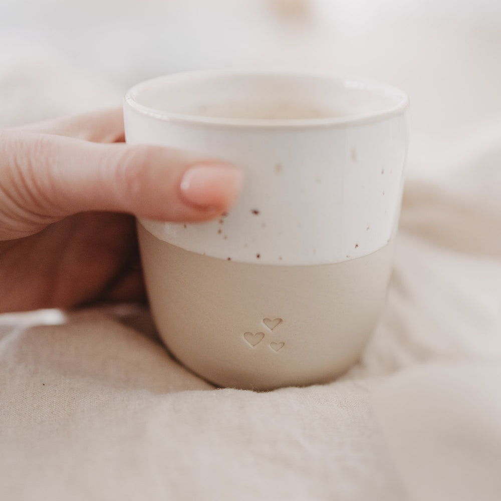 Beige-weißer Keramikbecher mit Herzprägung wird in der Hand gehalten – detailreiche Nahaufnahme.