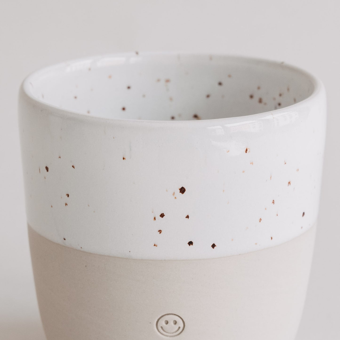 Keramikbecher mit weißer Sprenkelglasur und Smiley-Prägung, Fokus auf die glatte, helle Oberfläche.