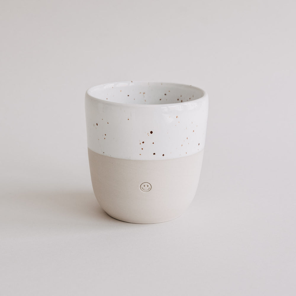 Heller Keramikbecher mit weißer Sprenkelglasur und eingeprägtem Smiley, freigestellt vor neutralem Hintergrund.