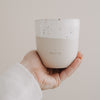 Hand hält beige-weiße „Ich lieb’s“-Tasse mit Sprenkelglasur vor hellem Hintergrund.