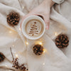 Blick in den Becher „O Tannenbaum“ mit Latte Art in Form eines Weihnachtsbaums, dekoriert mit Zapfen und Lichtern.