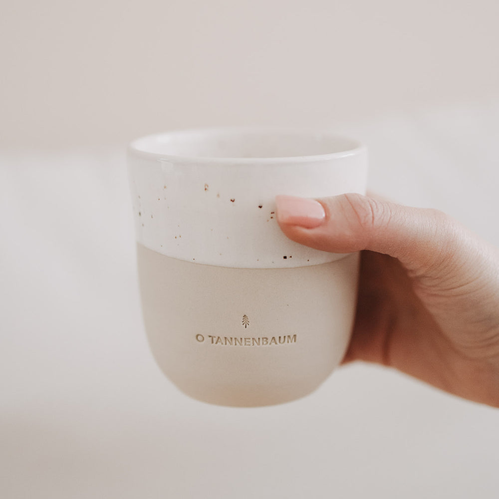 Hand hält den Becher „O Tannenbaum“ aus Keramik mit weißer Sprenkelglasur und matter beige-farbener Unterseite.