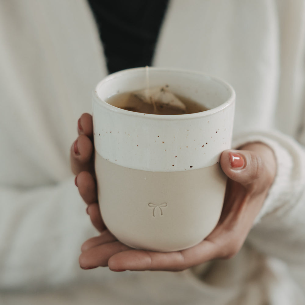 Keramikbecher mit Schleifenmotiv in Händen, gefüllt mit Tee, minimalistisches Design in warmem Licht
