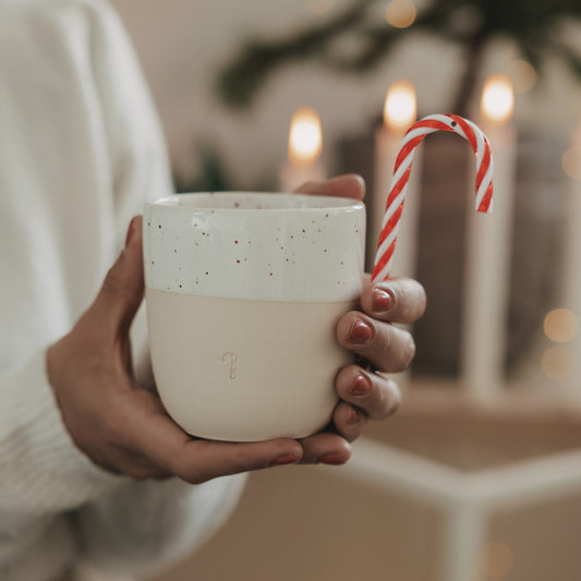 Keramikbecher mit Tee und Zuckerstange, in Händen gehalten, stimmungsvolle Weihnachtsdeko im Hintergrund