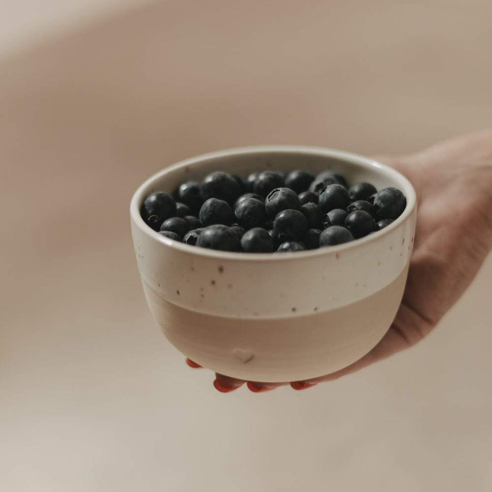 Beige-weiße Keramikschale mit Herz, gefüllt mit frischen Heidelbeeren und in der Hand gehalten
