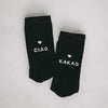 Schwarze Socken mit weißem Herzmotiv und den Aufschriften 'CIAO' und 'KAKAO', nebeneinander auf hellem Hintergrund liegend.