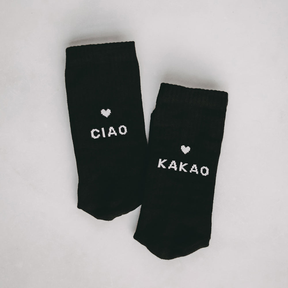 Schwarze Socken mit weißem Herzmotiv und den Aufschriften 'CIAO' und 'KAKAO', nebeneinander auf hellem Hintergrund liegend.