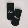Zwei schwarze Socken nebeneinander auf hellem Untergrund liegend, beide mit dem weißen Schriftzug "Moin" in Schreibschrift.