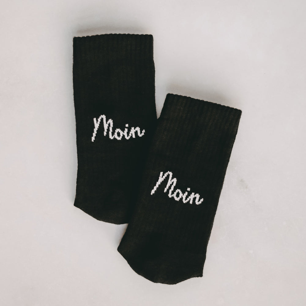 Zwei schwarze Socken nebeneinander auf hellem Untergrund liegend, beide mit dem weißen Schriftzug "Moin" in Schreibschrift.