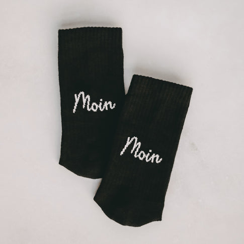 Zwei schwarze Socken nebeneinander auf hellem Untergrund liegend, beide mit dem weißen Schriftzug "Moin" in Schreibschrift.