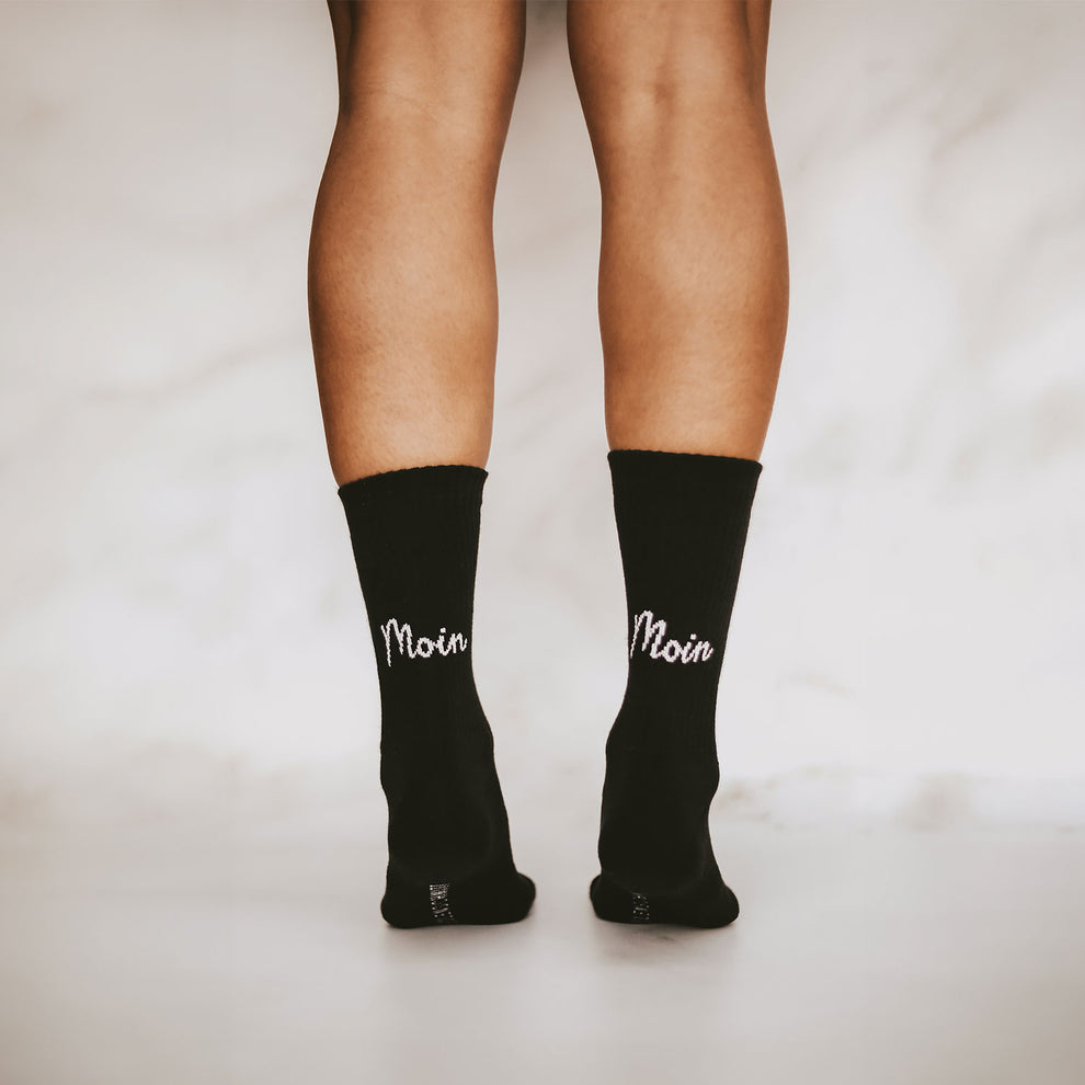 Rückansicht von zwei Beinen in schwarzen Socken mit weißem "Moin"-Schriftzug auf der Ferse; die Person steht auf hellem Boden vor hellem Hintergrund.