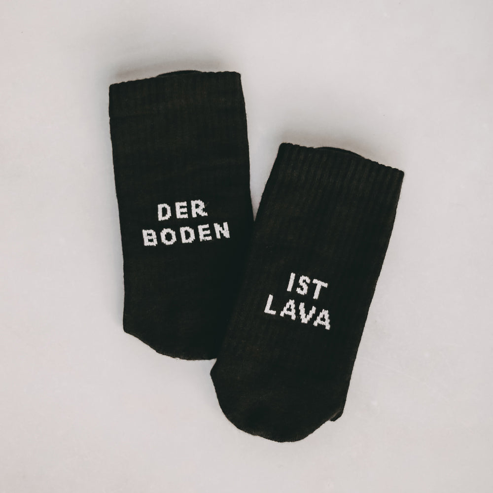 Zwei schwarze Socken liegen nebeneinander auf hellem Untergrund. Die linke Socke trägt den Schriftzug 'DER BODEN', die rechte 'IST LAVA'.