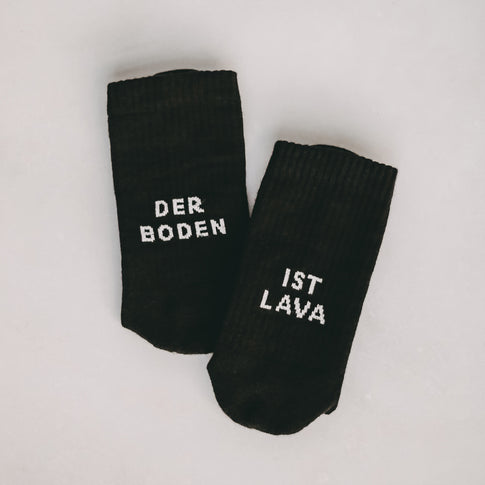 Zwei schwarze Socken liegen nebeneinander auf hellem Untergrund. Die linke Socke trägt den Schriftzug 'DER BODEN', die rechte 'IST LAVA'.
