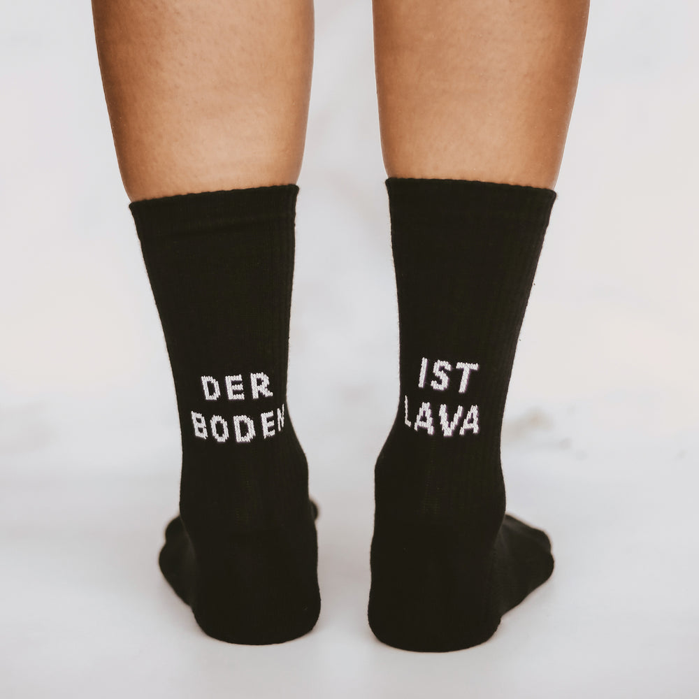 Nahaufnahme von zwei Beinen in schwarzen Socken mit weißer Schrift. Auf der linken Socke steht 'DER BODEN', auf der rechten 'IST LAVA'.