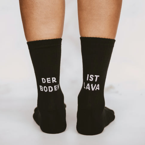 Nahaufnahme von zwei Beinen in schwarzen Socken mit weißer Schrift. Auf der linken Socke steht 'DER BODEN', auf der rechten 'IST LAVA'.