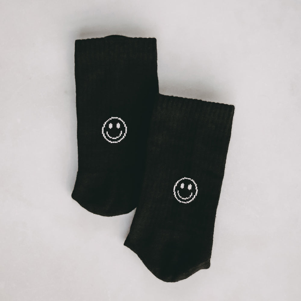 Schwarze Socken mit weißem Smiley-Motiv auf hellem Untergrund flach nebeneinander gelegt.