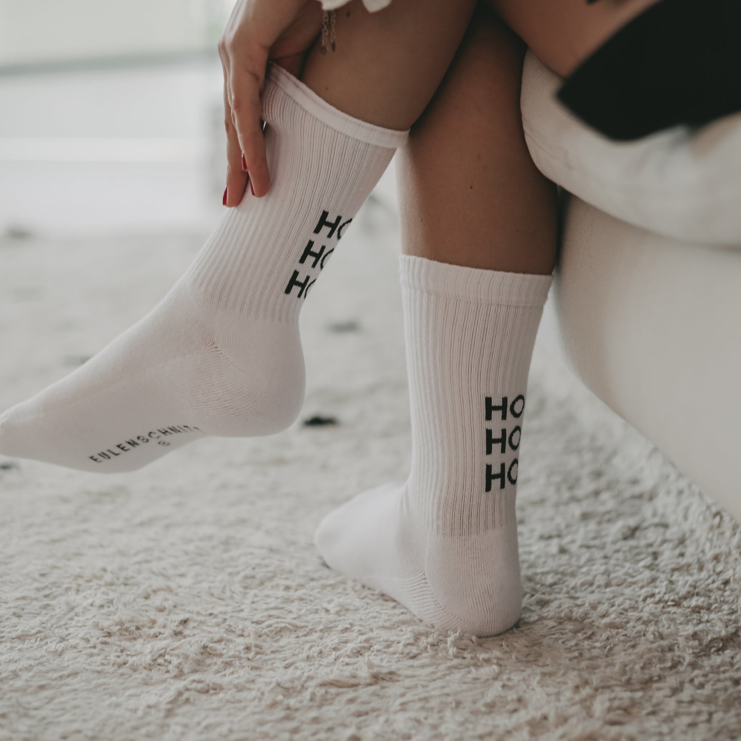 Nahaufnahme von Beinen mit weißen Socken mit schwarzem ‚HO HO HO‘ Schriftzug, sitzend auf einem Sofa mit beigem Teppich.