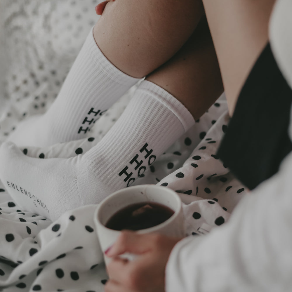 Nahaufnahme von Beinen mit weißen Socken mit schwarzem Schriftzug ‚HO HO HO‘, eine Hand hält eine Tasse Tee. Die Person sitzt gemütlich auf einem Bett mit gepunkteter Decke.
