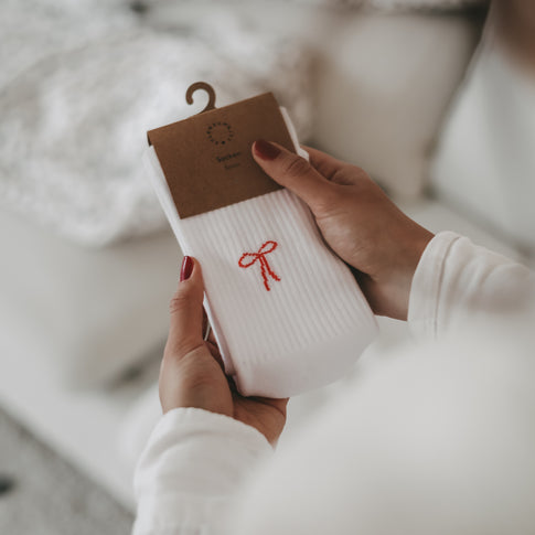 Nahaufnahme von Händen, die ein Paar weiße Socken mit rotem Schleifen-Motiv in einer Kraftpapier-Verpackung mit Aufhängung halten.