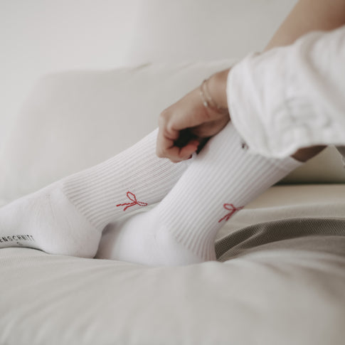 Frau sitzt auf einem Sofa, Nahaufnahme von Beinen in weißen Socken mit roter Schleife, eine Hand berührt die Socke.