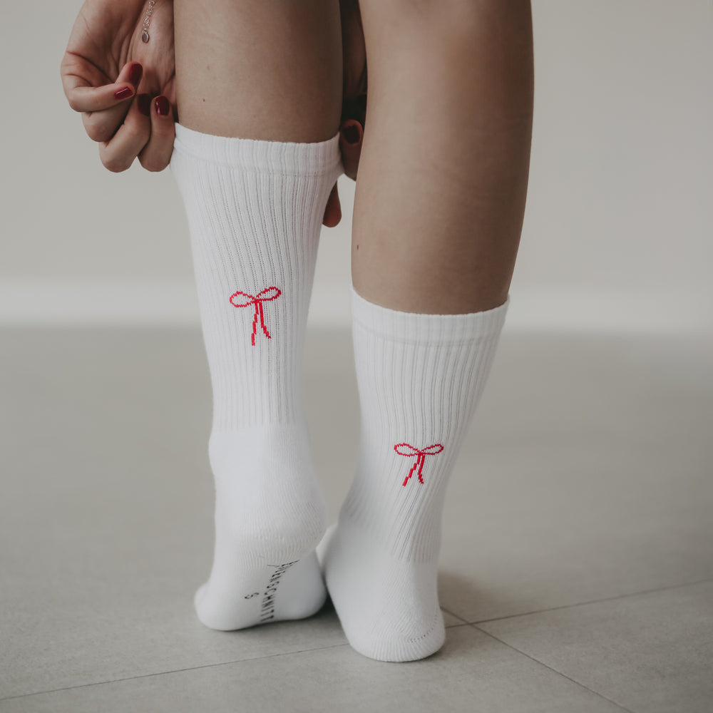 Nahaufnahme: Frau steht auf hellem Boden, trägt weiße Socken mit rotem Schleifen-Motiv auf den Fersen und fasst sich mit beiden Händen an die linke Wade.