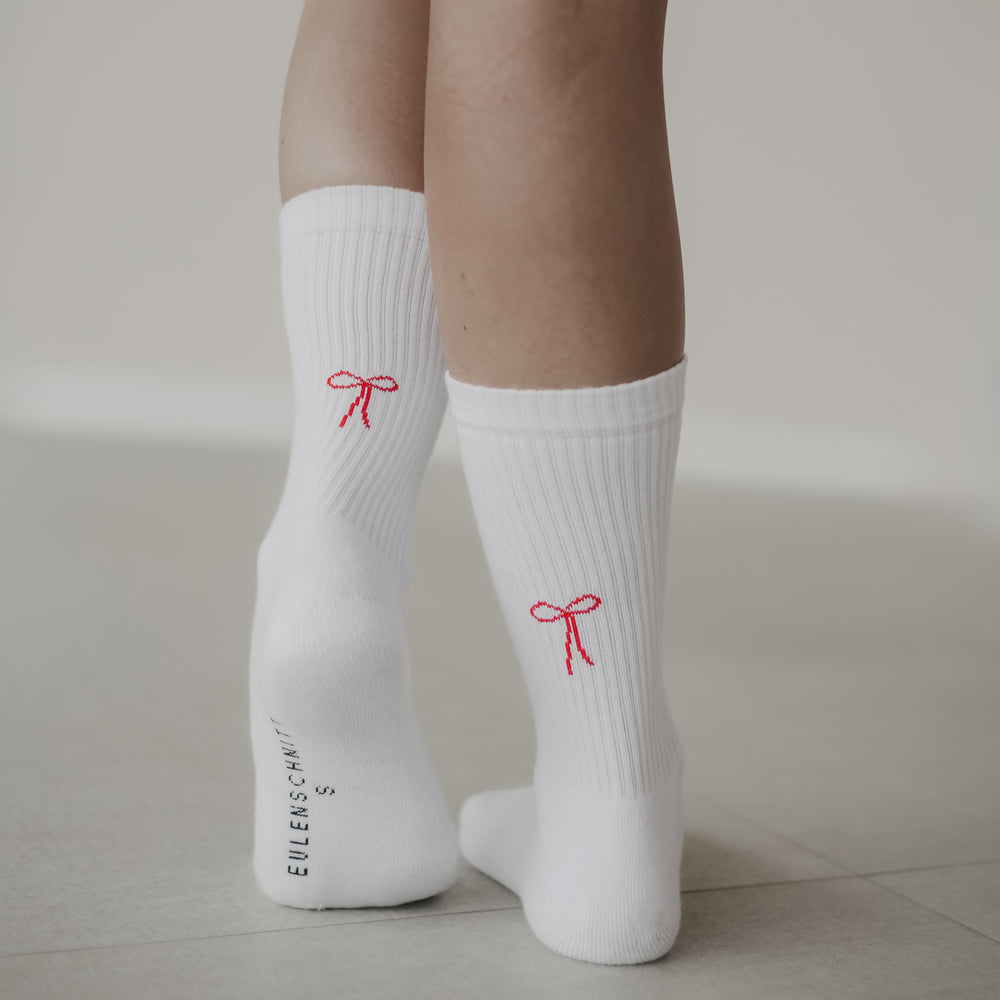 Nahaufnahme: Frau steht auf hellem Boden, trägt weiße Socken mit rotem Schleifen-Motiv auf den Fersen.