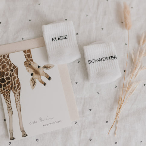 Babysocken „Kleine Schwester“ neben einer Giraffenkarte und Trockenblumen auf hellem Stoff.