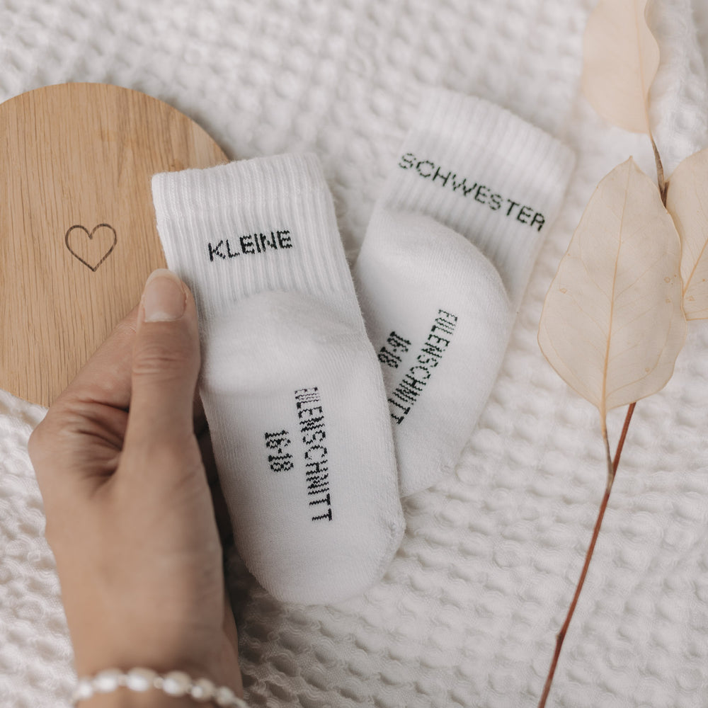 Hand hält Babysocken „Kleine Schwester“ auf strukturierter Decke mit Holzuntersetzer und Blättern.