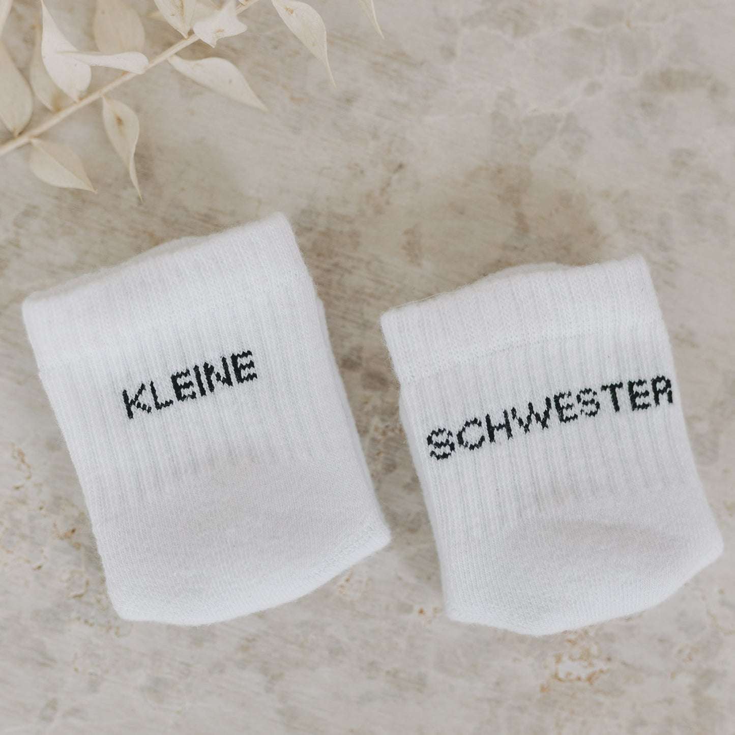 Babysocken „Kleine Schwester“ auf strukturierter  Unterlage und Blättern.