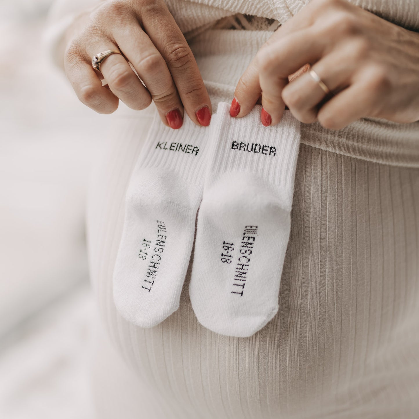 Hände halten Babysocken „Kleiner Bruder“ auf Babybauch vor heller Kulisse