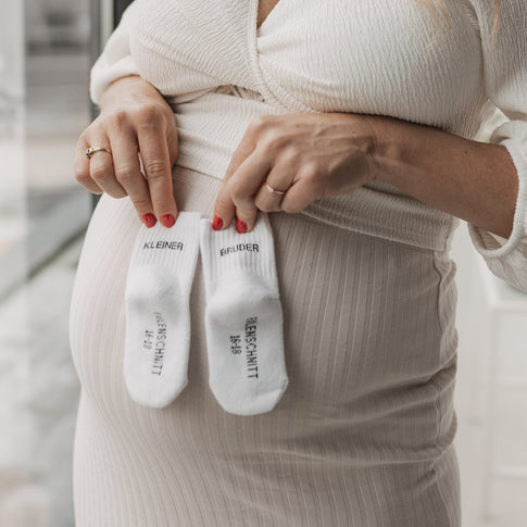 Schwangere zeigt Babysocken „Kleiner Bruder“ vor dem Babybauch