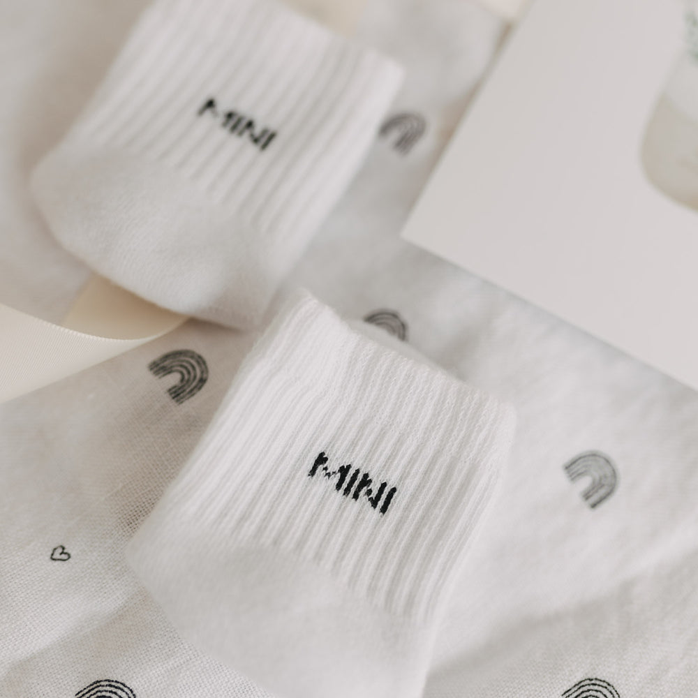 Detailaufnahme der weißen „MINI“-Socken auf Tuch mit Regenbogenmotiv und Herzsymbolen