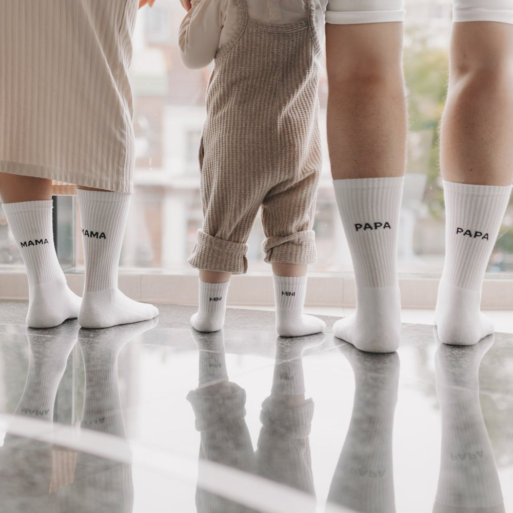 Familie trägt passende Socken mit „MAMA“, „PAPA“ und „MINI“-Schriftzug, steht gemeinsam am Fenster