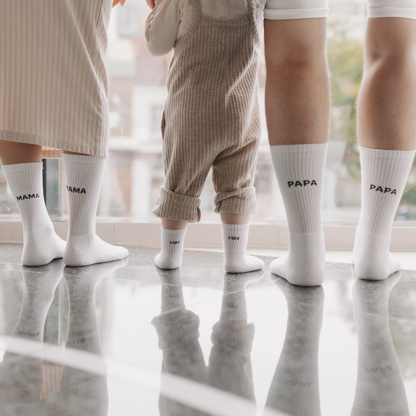Familie trägt passende Socken mit „MAMA“, „PAPA“ und „MINI“-Schriftzug, steht gemeinsam am Fenster