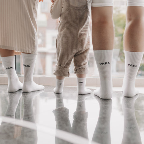 Familie trägt passende Socken mit „MAMA“, „PAPA“ und „MINI“-Schriftzug, steht gemeinsam am Fenster