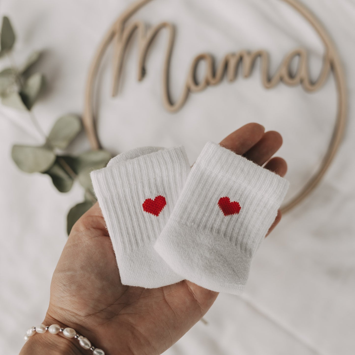 Zwei Babysocken mit rotem Herz in einer Hand gehalten, im Hintergrund ein „Mama“-Dekoelement aus Holz und Eukalyptus.