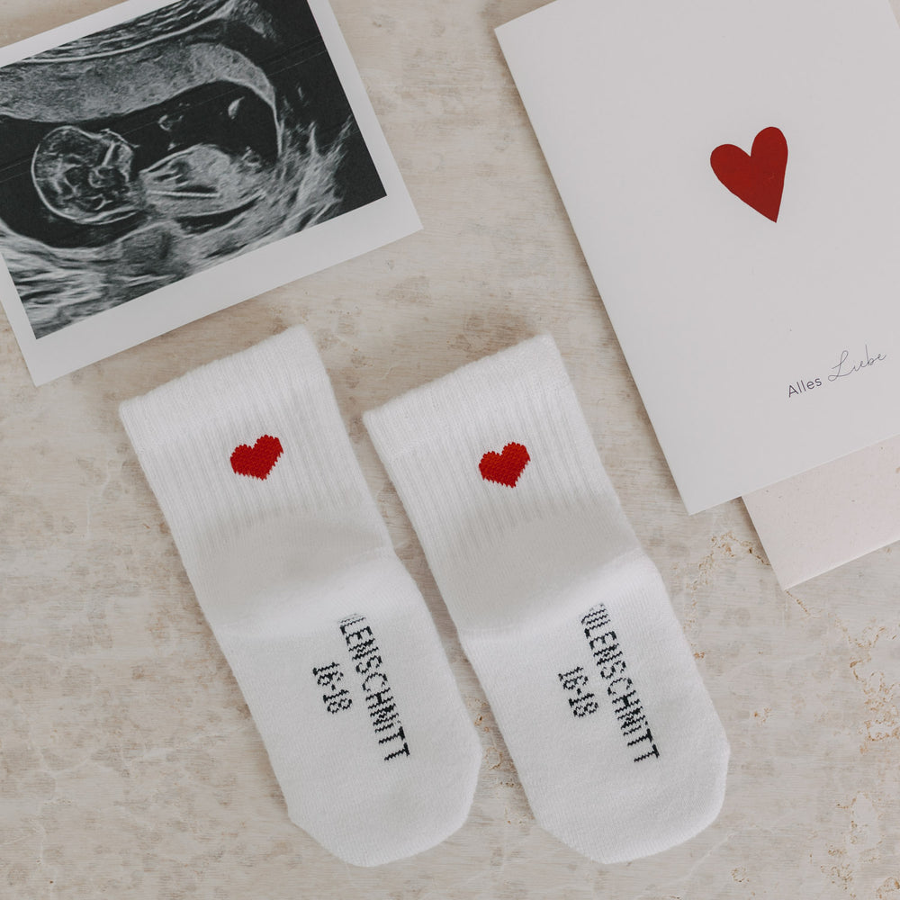 Babysocken mit rotem Herz neben Ultraschallbild und Karte mit Herzmotiv – liebevolle Geschenkidee zur Geburt.