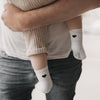 Baby trägt weiße Socken mit schwarzem Herzmotiv, gehalten von einem Erwachsenen in Jeans und T-Shirt.