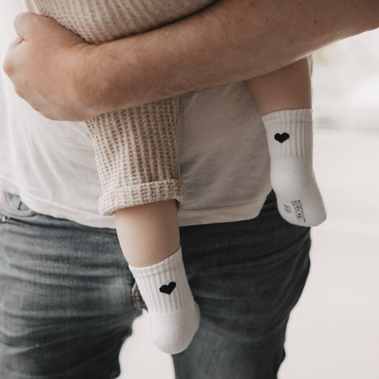 Baby trägt weiße Socken mit schwarzem Herzmotiv, gehalten von einem Erwachsenen in Jeans und T-Shirt.