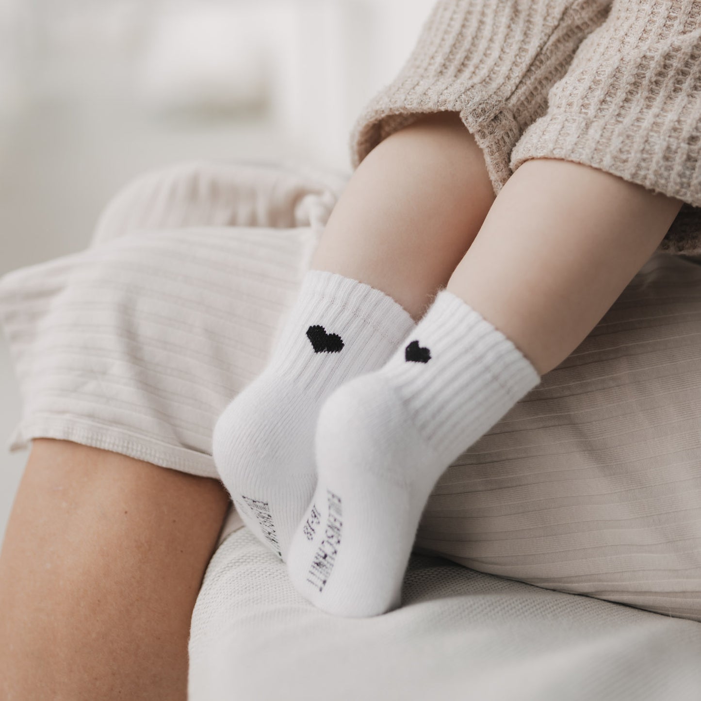 Baby sitzt auf Schoß mit weißen Socken mit schwarzem Herz, Füße im Fokus vor heller, weicher Umgebung