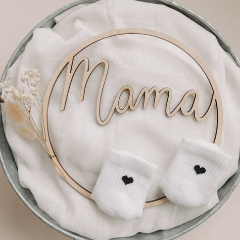 Zwei Babysocken mit schwarzem Herz auf weißem Stoff, dekorativ mit Holzschriftzug „Mama“ arrangiert.