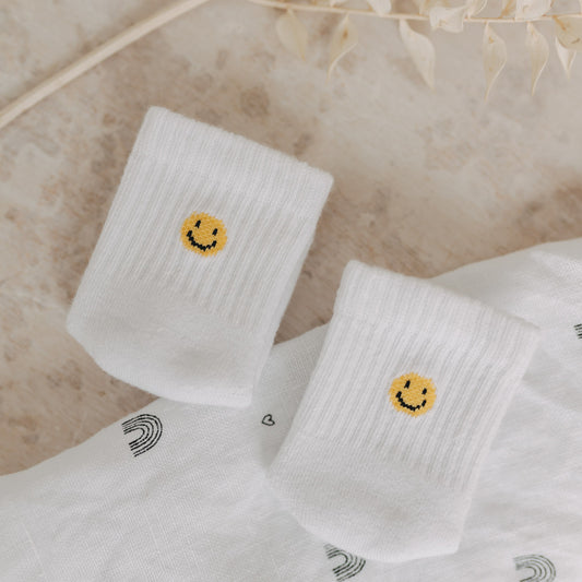 Weiße Babysocken mit gelbem Smiley auf gemustertem Stoff, dekoriert mit Trockenblumen.