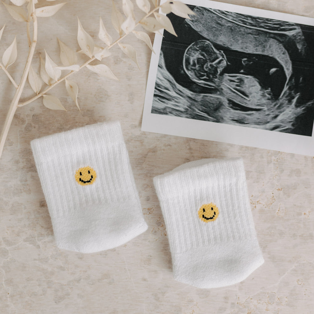 Smiley-Babysocken neben einem Ultraschallbild und Trockenblumen auf hellem Untergrund.