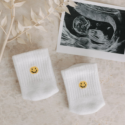 Smiley-Babysocken neben einem Ultraschallbild und Trockenblumen auf hellem Untergrund.