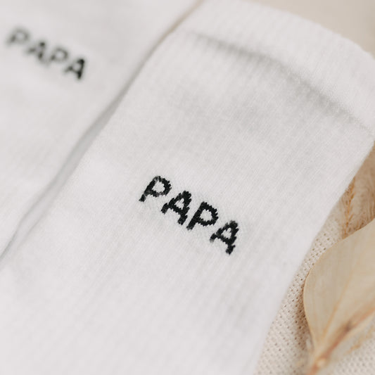 Nahaufnahme des schwarzen Schriftzugs „PAPA“ auf weißen Socken mit feiner Struktur.