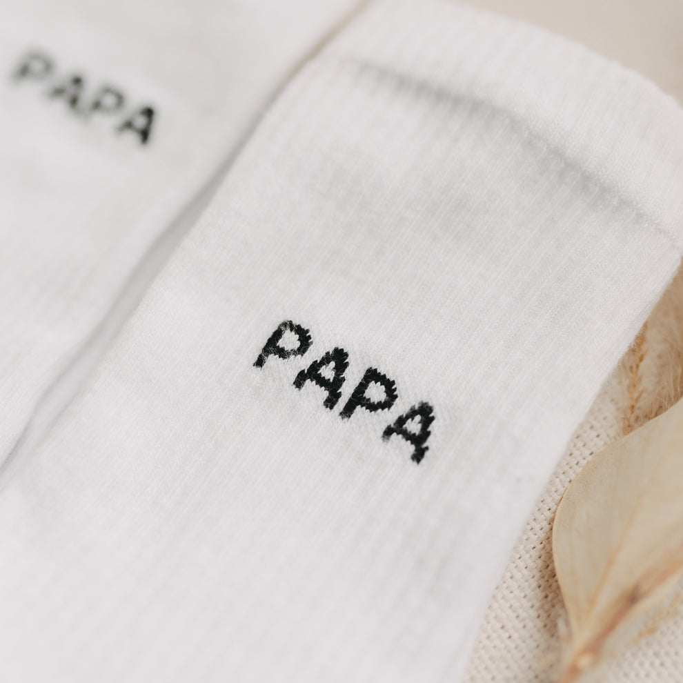 Nahaufnahme des schwarzen Schriftzugs „PAPA“ auf weißen Socken mit feiner Struktur.