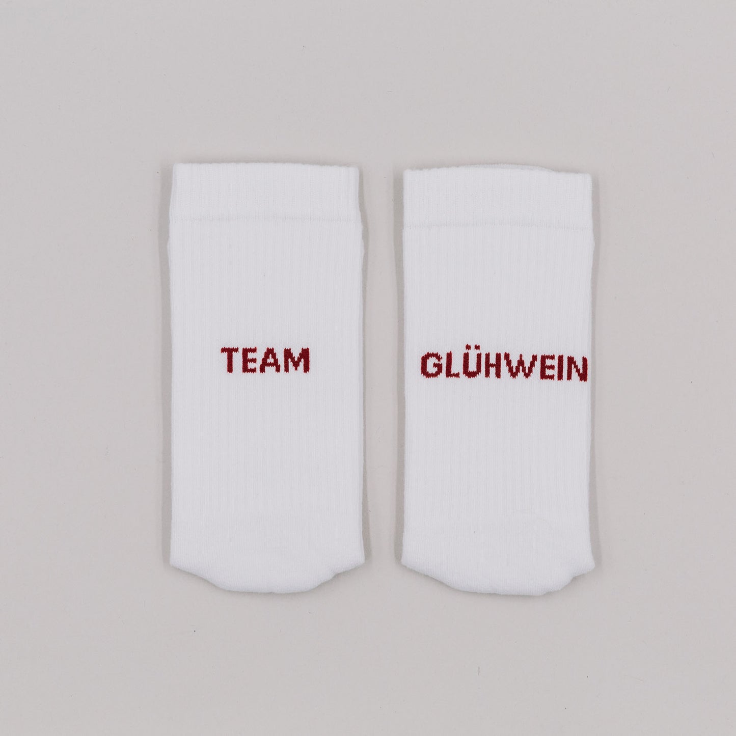 Weiße Socken mit rotem Schriftzug „Team Glühwein“ nebeneinander liegend vor neutralem Hintergrund.
